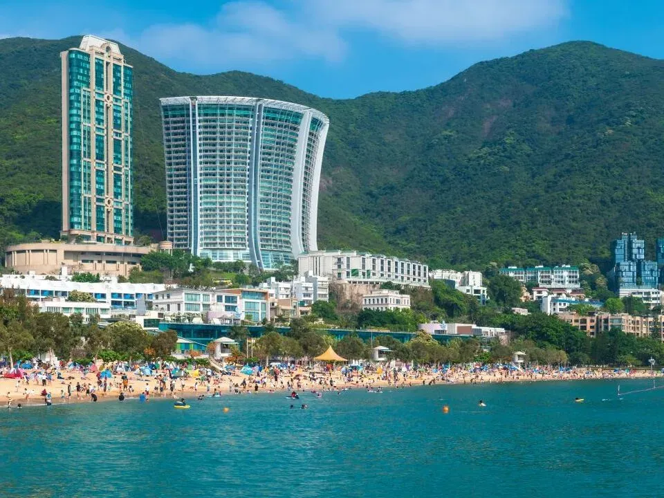 浅水湾的阳光明媚海滩景观，人群熙攘、碧绿海水与现代摩天大楼。
